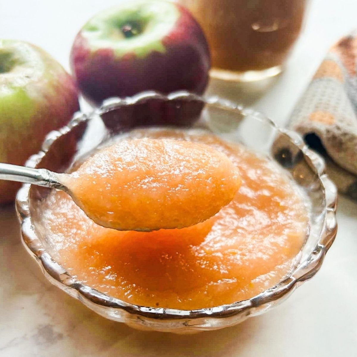 bowl of applesauce with spoon holding applesauce and 2 apples behind it on table.