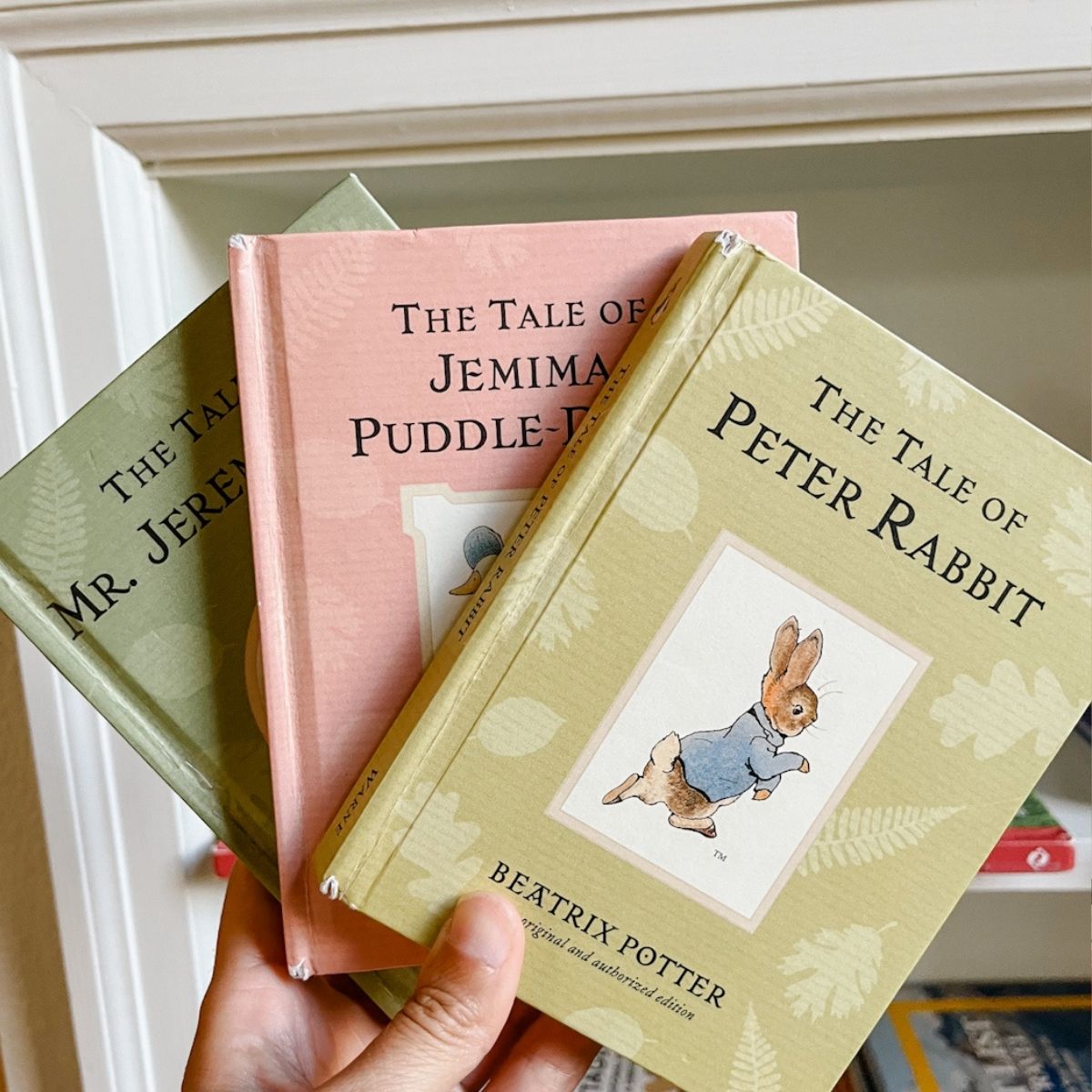 Three whimsical books by Beatrix Potter held up in front of a bookshelf.