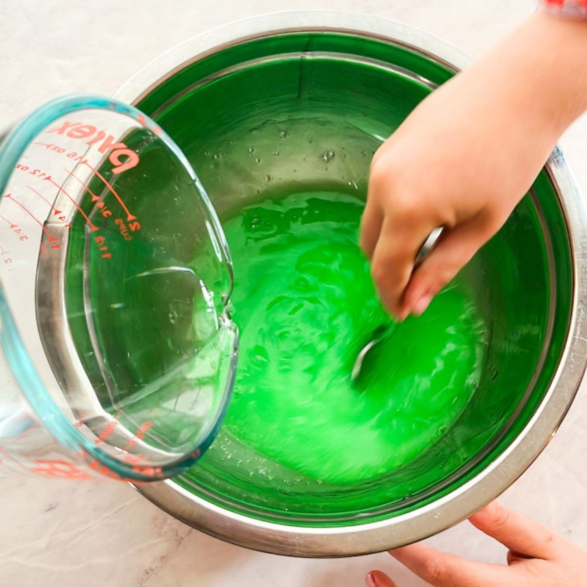 Measuring cup pouring borax solution into the large mixing bowl while a hand is stirring it.